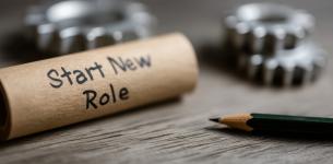 Image of a sign stating 'start new role' placed on a table beside a pencil and metal cogs.
