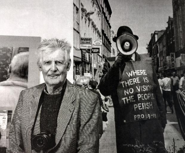 Black and white photograph of a man with a camera around his neck standing in front of a photographic exhibition