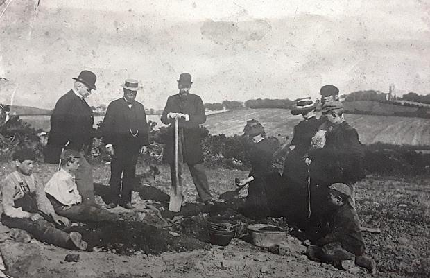 Old black and white photograph showing 10 individuals and a small dog at a archaeological excavation
